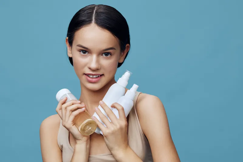 Young girl holding skincare products on a blue background, promoting healthy skin and beauty routines for all ages