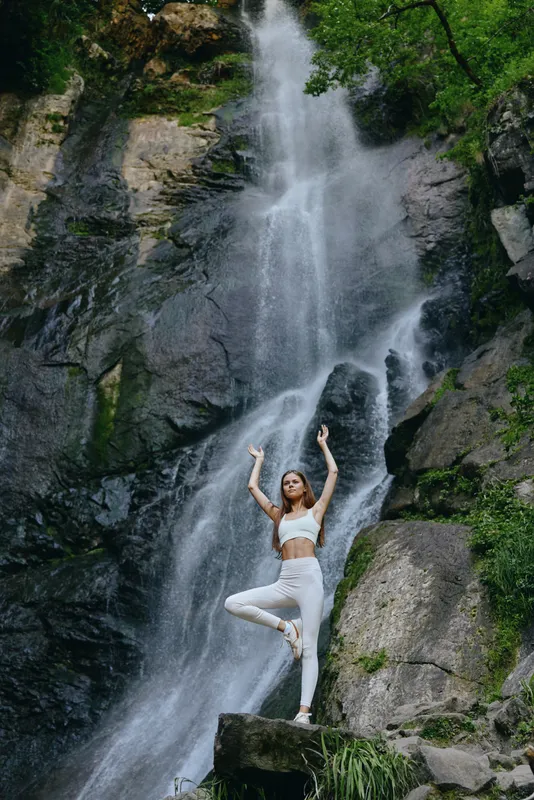 woman practicing yoga poses near a high waterfall in nature, wearing white sportswear and surrounded by lush green rocks and flowing water
