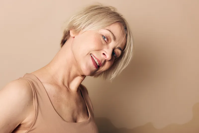 Smiling mature woman with short blonde hair wearing a beige tank top poses against a neutral background. Concept of natural beauty, confidence, and aging gracefully in lifestyle portrait.