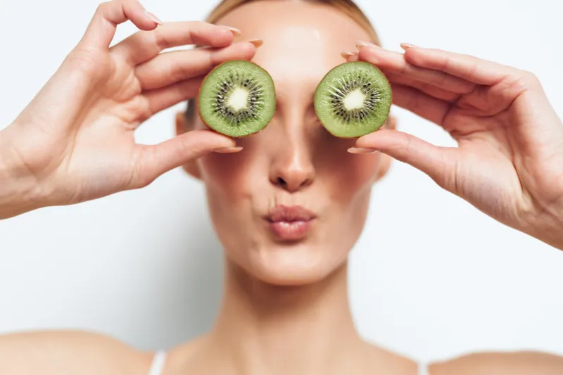 A playful young woman holds two kiwi halves over her eyes, puckers her lips, and gazes at the camera in a bright studio, creating a lighthearted wellness moment.