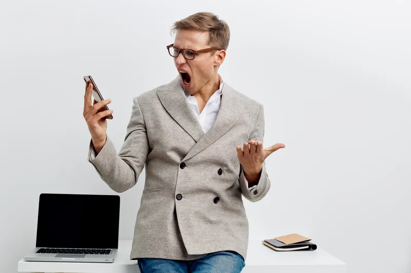 frustrated man using smartphone in modern blazer eyeglasses standing near laptop white background yelling angry digital detox concept technology stress