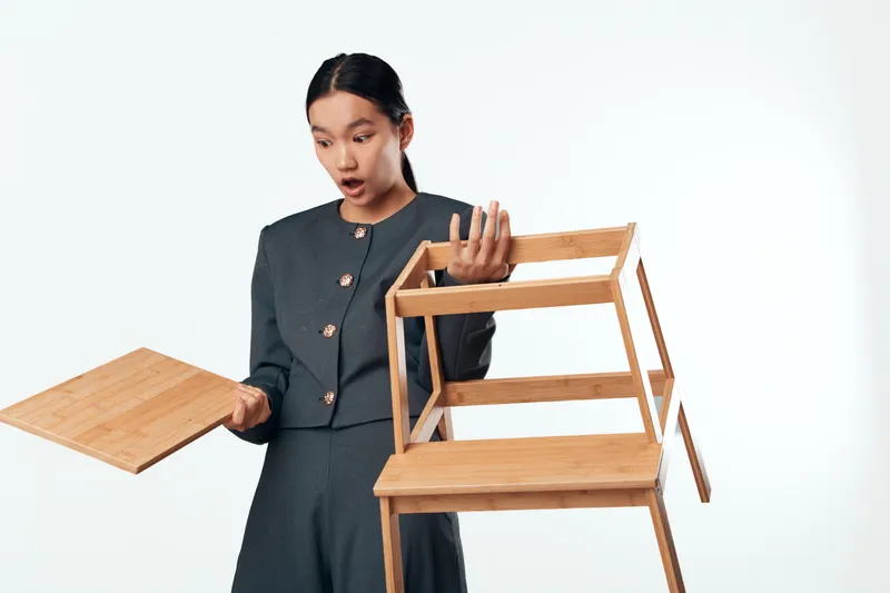 Woman in gray suit assembling wooden furniture holding chair frame and panel, surprised expression on face, isolated on white background, woodworking and DIY project concept.