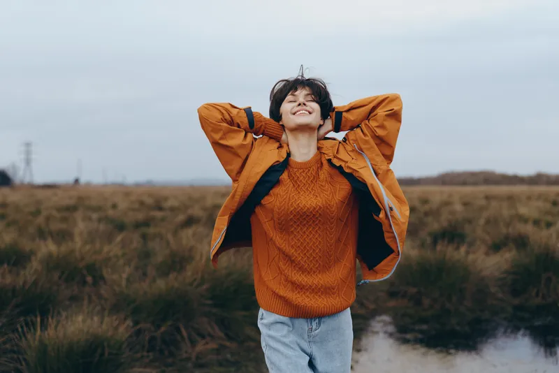Woman, lifestyle, outdoor, field, autumn, windy, sweater, jeans, nature, smiling, standing on a rural path, hands behind head, looking up, soft daylight, casual clothing, peaceful seasonal atmosphere