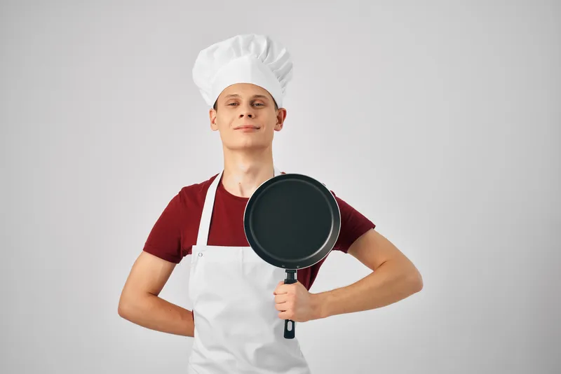 Повар в профессиональной униформе держит в руках сковороду на кухне