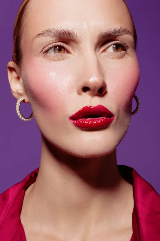 Close-up of a woman with glossy red lips and natural skin wearing hoop earrings and a red top against a purple background. Concept of beauty, makeup, and fashion portrait.