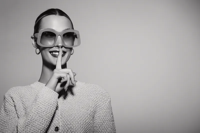 Stylish young Caucasian woman with fair skin wearing sunglasses and a sweater makes a shushing gesture with her finger.