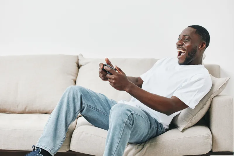 Happy Black man enjoying gaming on the sofa, smiling joyfully while playing video games in casual attire with a modern minimalist background