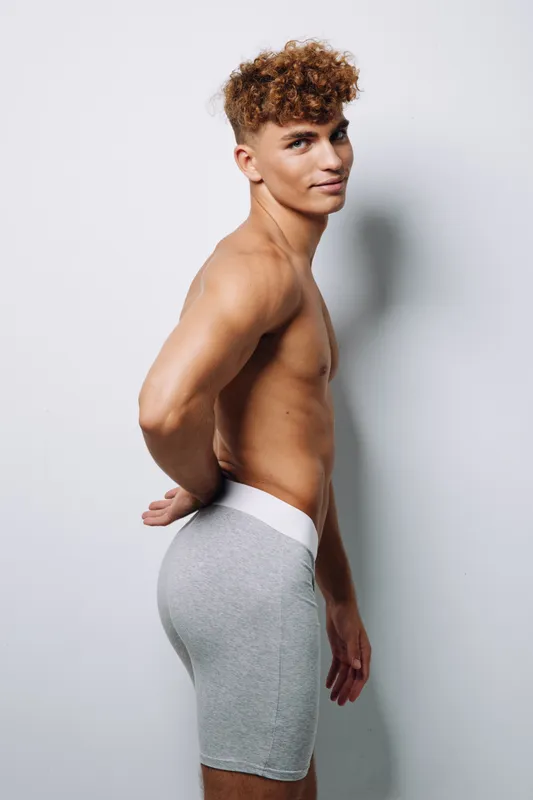 Young man with curly hair standing sideways against white background in gray shorts, healthy slim body, and relaxed confident expression with hands behind back in studio.