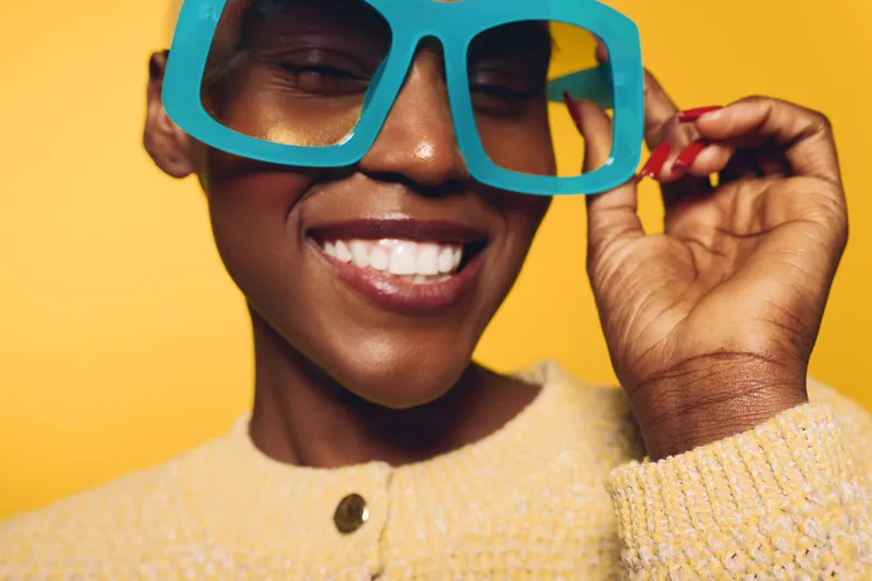 Close-up of smiling African woman with dark skin wearing oversized blue glasses and yellow knit sweater against yellow background. Concept of fashion, joy, and vibrant style in studio.