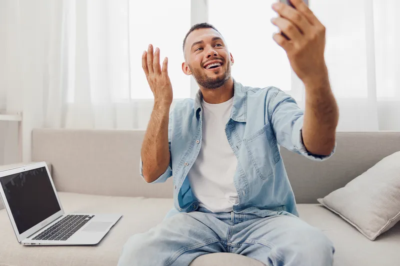 Smiling man engaged in a video call, expressing joy. Bright and airy living room setting, showcasing modern lifestyle and connection through technology.