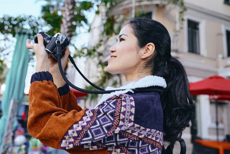 Camera in hand, woman photographer gazes upward as she captures a candid street scene outdoors, wearing a patterned sweater and jacket that add color to urban architecture in daylight