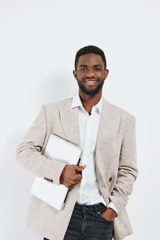 Confident young African man in smart beige blazer and white shirt holding a laptop and documents, smiling, standing against a plain white background, studio portrait. Business professional concept