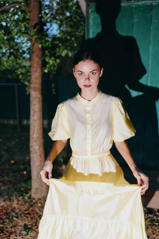 Woman in a yellow dress stands outdoors in daylight, a bold shadow on a wall behind her, creating a vintage fashion portrait with natural sunlit light