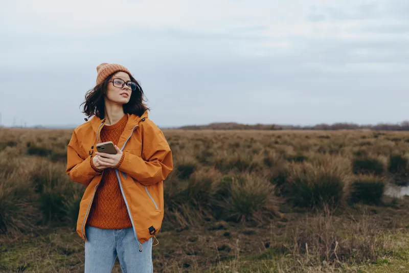 Woman lifestyle outdoors, casual autumn field, hat and jacket, holding phone, relaxed smile, cloudy sky, natural light, grassy landscape, soft breeze, quiet nature walking, calm moment