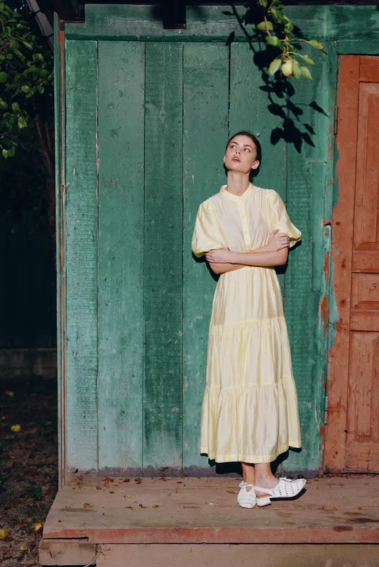 Elegant woman in a pale yellow dress leans against a rustic green door, posing thoughtfully in an outdoor setting with soft light, calm mood, and simple surroundings