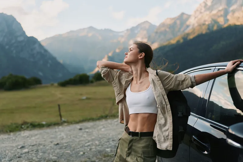 A woman stands beside a car with arms outstretched, embracing mountain air. Casual outfit, white crop top and rugged jacket. Concept of freedom and outdoor lifestyle.