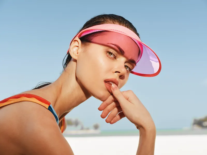 Close-up portrait of a stylish woman wearing a pink visor at a sunny beach, sunlit skin, relaxed pose, confident, casual summer vibe today.