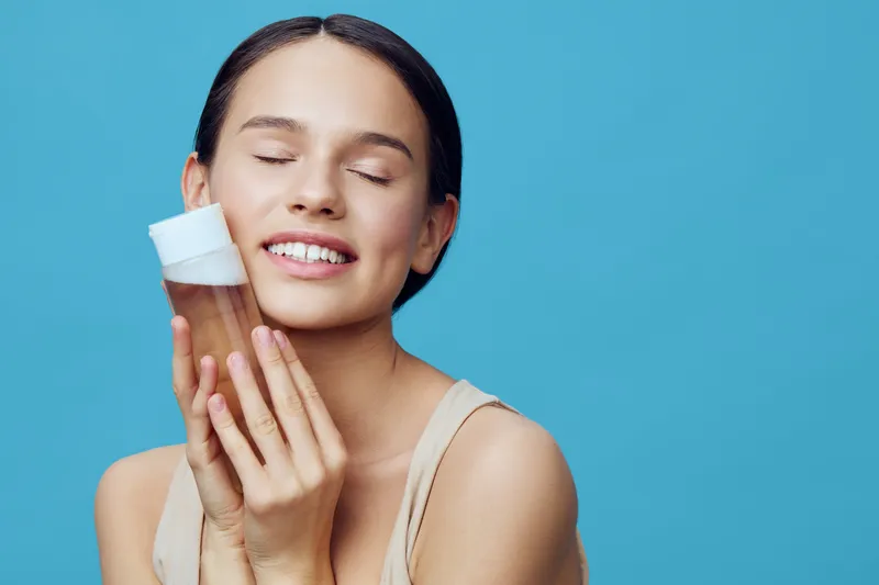 Young woman smiling with a skincare product against a blue background, showcasing healthy skin and beauty care routine