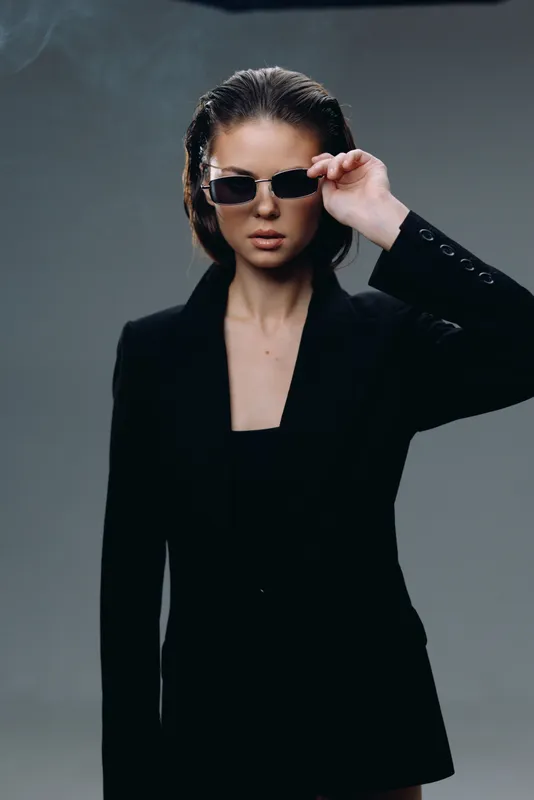 Stylish woman wearing black blazer and sunglasses with confident expression against gray background