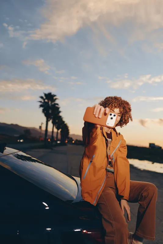 Woman lifestyle outdoors at sunset, curly hair in orange jacket, sitting by car, holding teddy dog close, playful mood, soft evening light, calm street background