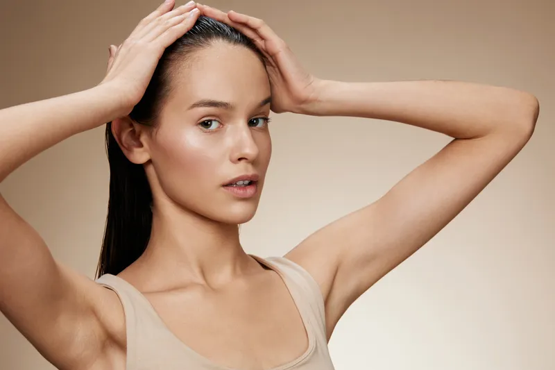 Young woman with long hair holding her head, showcasing healthy hair and skin on a neutral background, exuding calmness and confidence