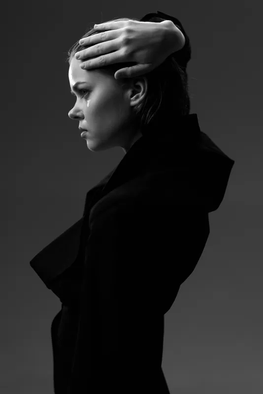 Black and white portrait of a thoughtful woman with hand on head in dark clothing against dark background.