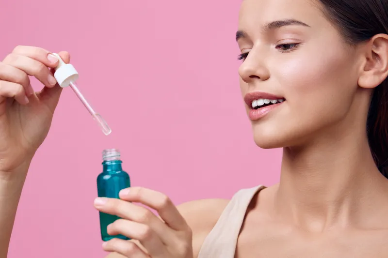 Young woman with natural beauty applying serum from a dropper bottle against a pastel pink background, conveying a sense of skincare and self care routines