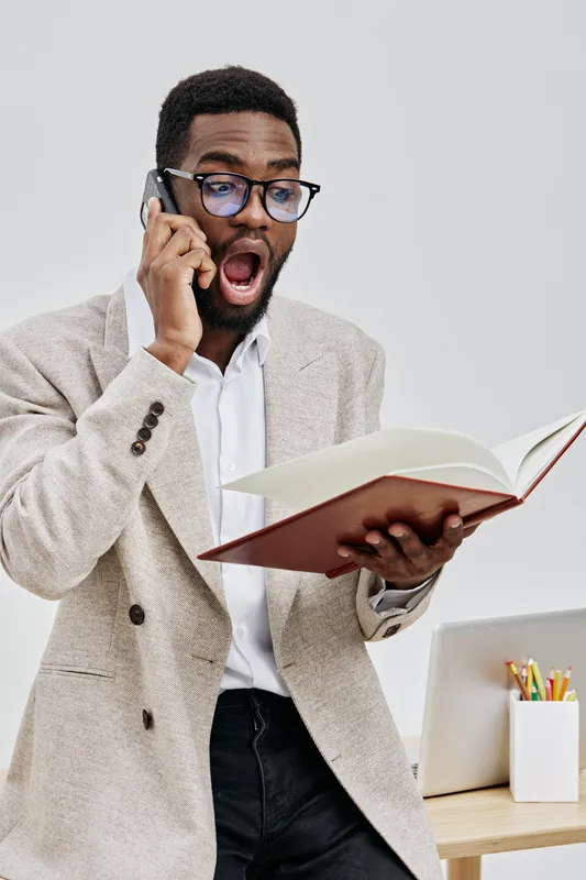 Excited surprised man talking on mobile phone while reading a book in office environment. Young African American businessman wearing glasses and beige blazer expressing astonishment. People lifestyle 