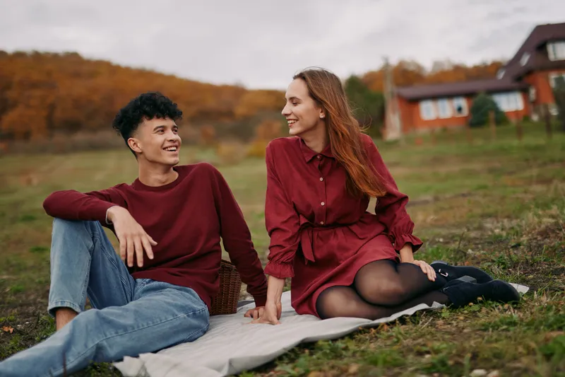 A real moment between a couple outdoors on autumn grass, warm smiles, natural light, simple wardrobe, genuine connection, and a calm, candid mood.
