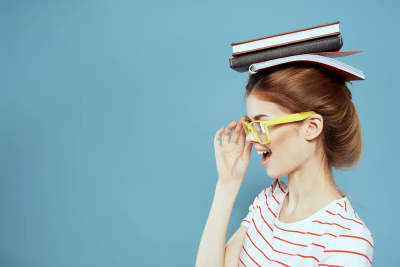 Студентка держит книги на голове и в жёлтых очках на синем фоне, обрезанный вид