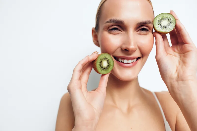 Smiling young woman holds two halves of fresh kiwi fruit near face isolated on white background healthy lifestyle concept natural skincare and organic nutrition