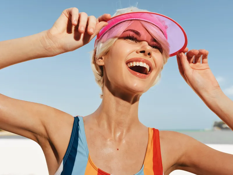 Joyful woman on a sunny beach adjusts her pink visor, laughing with bright teeth and radiant skin, capturing carefree summer energy and athletic confidence under clear blue skies.