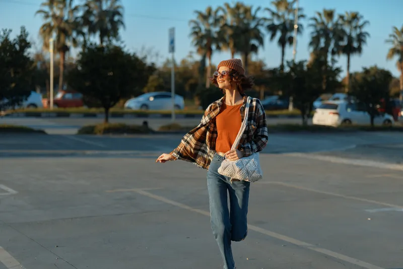 Woman walking in golden sunlight across a parking lot, candid moment of authenticity and lifestyle captured with a smile, casual bohemian outfit and relaxed urban evening glow.