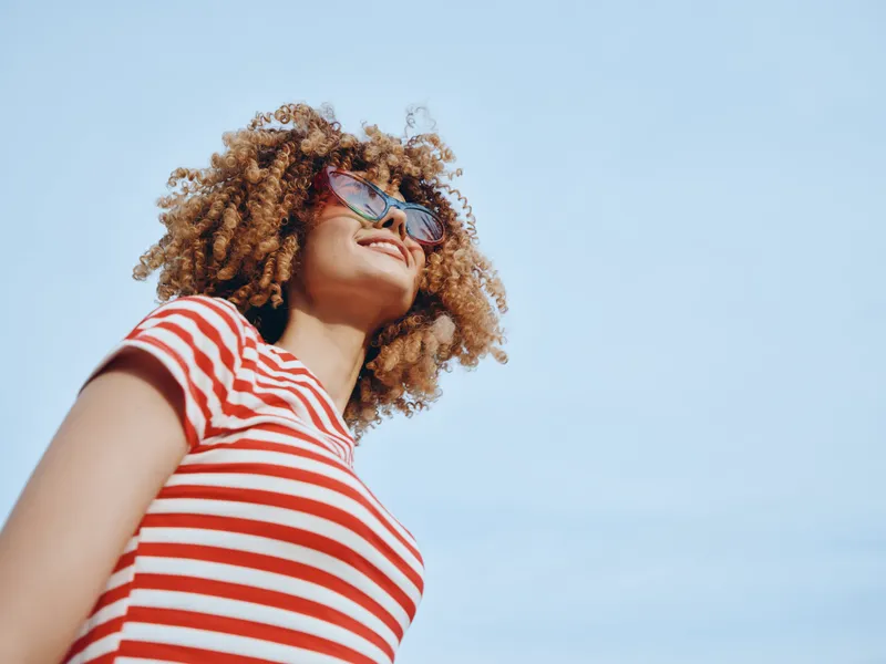 Portrait of a joyful young person with curly hair, wearing a red and white striped shirt and sunglasses, standing outdoors against a clear blue sky.