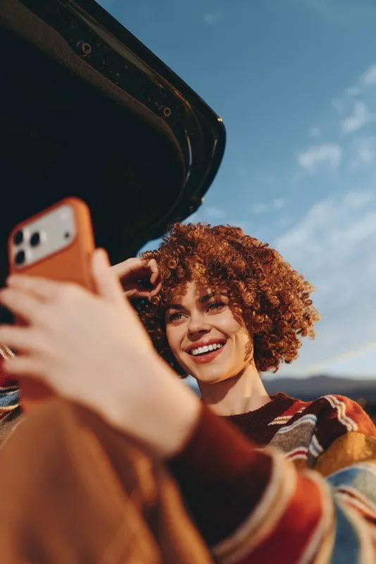 Woman with curly hair smiles in outdoor lifestyle selfie, phone near face against blue sky and mountains, warm golden light, casual travel mood from scenic viewpoint by the sea today.