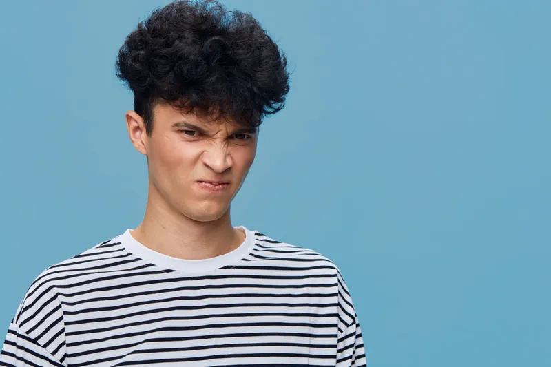 Young man with curly hair making a disgusted face wearing a striped shirt against a solid blue background, showing emotion and casual style.