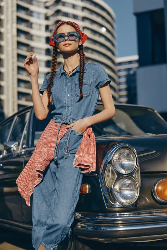 modern boho-western denim street style woman with sunglasses and braided hair posing by vintage car under city sunlight in film color mood