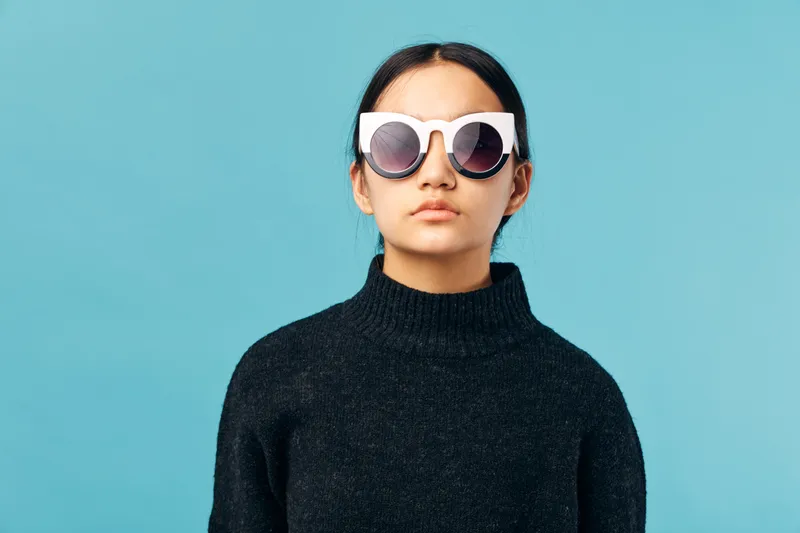 Young woman wearing large white sunglasses and black sweater stands against blue background with neutral expression and straight dark hair for fashion and style.