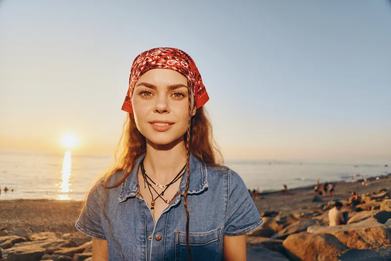 Lifestyle woman in modern boho-western denim street style wearing a red bandana, standing by the beach at sunset with warm film color tones enhancing natural light.