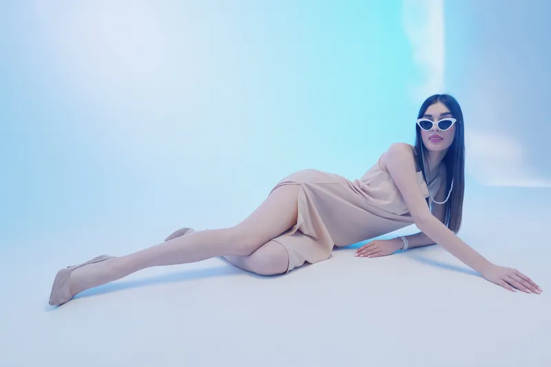 Elegant woman in a beige dress and sunglasses poses on the floor in a bright studio. Concept of beauty, fashion, style, and modern lifestyle.