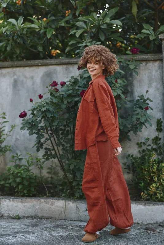 Cheerful woman lifestyle outdoors, wearing rust orange oversized suit in a garden, relaxed pose, soft light, natural colors, candid moment, curly hair frame