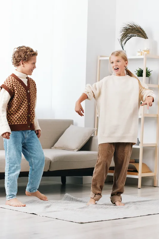 Two children dance joyfully in a bright, cozy living room, wearing soft sweaters as they move with wide smiles, capturing playful energy and carefree childhood in a candid moment.