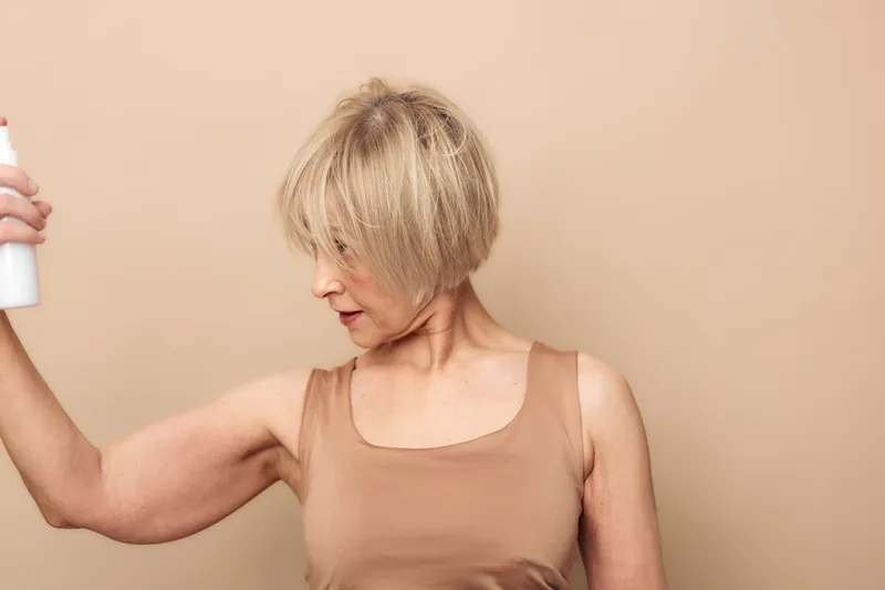 Mature woman with short blonde hair sprays deodorant on her armpit in a beige tank top against a plain neutral background. Concept for personal care, hygiene, and confidence.