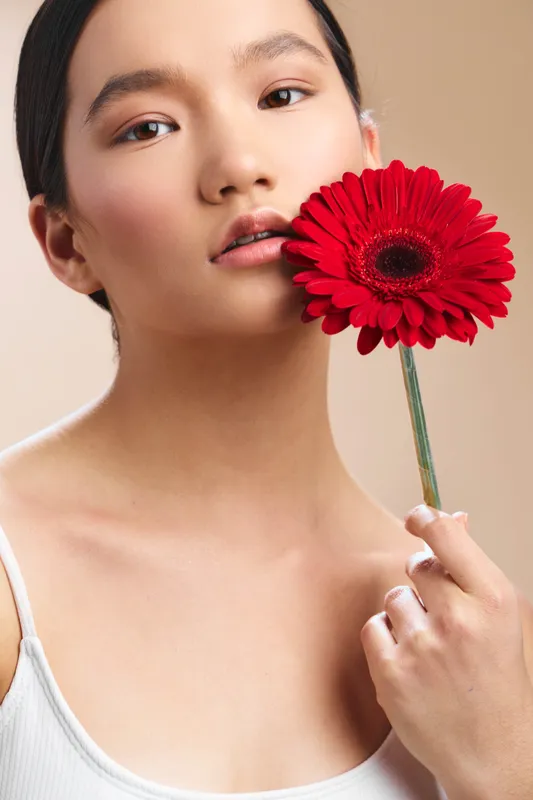 asian woman holds a bright red gerbera flower near her face in a soft studio. The portrait emphasizes natural beauty and delicate features, creating a calm, intimate mood with a bold floral focal