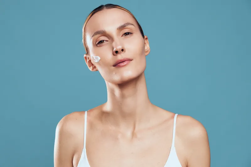 Smiling woman with a dab of white cream on her cheek, closeup portrait against a blue background. Focus on skincare, selfcare, and beauty routine with soft lighting