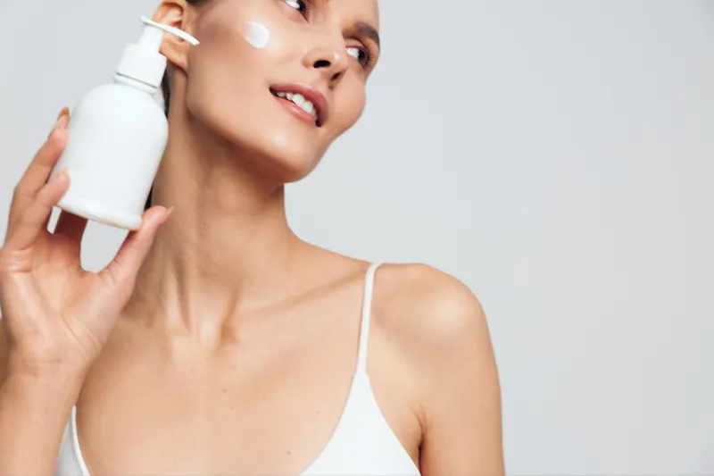 Young woman with clear skin applies moisturizing cream on her face holds white lotion bottle looks up with gentle smile isolated on light background skincare concept