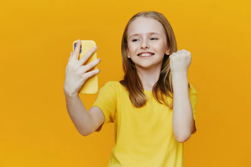 Young girl celebrating success while taking a selfie against a vibrant yellow background, showcasing joy and positivity in a casual yellow T shirt