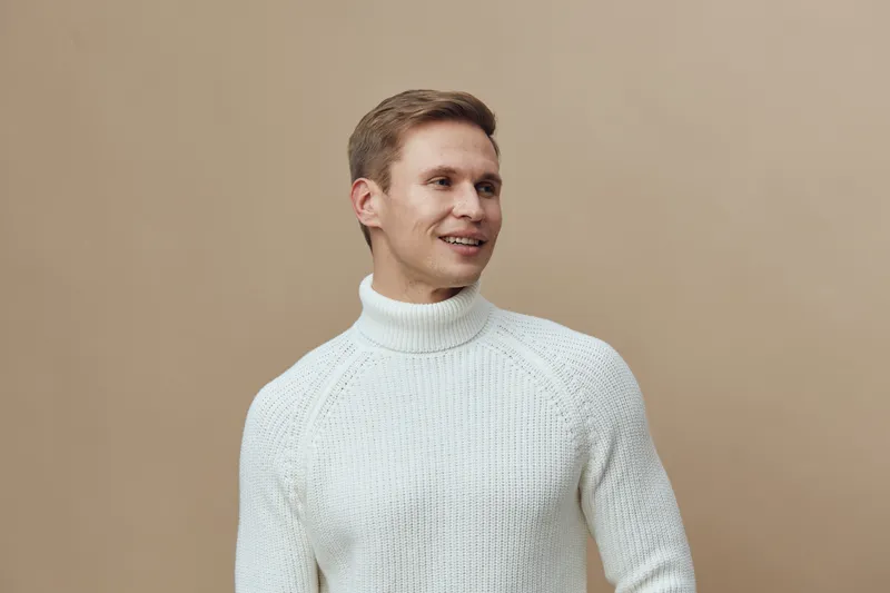 Smiling young caucasian man wear white knitted turtleneck sweater look side positive emotion with beige background lifestyle portrait mental health gadgets concept