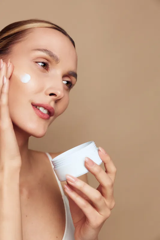 Young woman applying moisturizing cream on face, skincare routine, natural beauty, smooth skin, beige background, close-up portrait, gentle touch, wellness concept