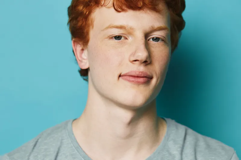 Young man with red hair and fair skin wearing a gray shirt poses against blue background looking confidently at camera with slight smile in studio portrait lighting.